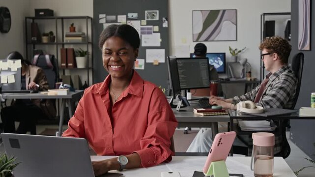 Portrait of African American young woman typing on laptop, turning to smile at camera in IT office. Footage for software development and diversity in tech promotions