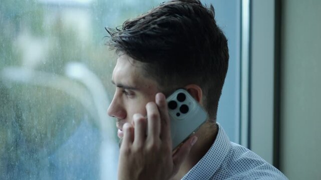 Portrait of male entrepreneur in shirt speaking by smartphone near large window into skyscraper. Young confident businessman talk on phone at office. Handsome manager having conversation on cellphone