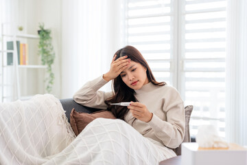 Fototapeta premium Unhealthy Woman feeling unwell, hand touching forehead, holding fever thermometer for measure temperature, resting on sofa at home. Health care.