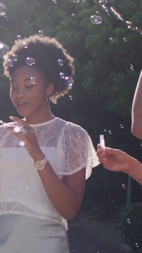 Vertical video: Blowing woman in lace top after friend dipping wand in garden, playing with bubbles