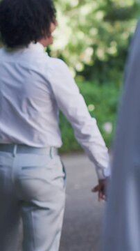 Vertical video: Panning couple holding hands walking to ceremony along garden path in white shirts