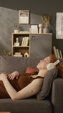 Vertical shot of pregnant Caucasian young adult woman with glucose monitor lying on sofa and listening to music. Perfect for marketing campaigns on gestational diabetes and prenatal care