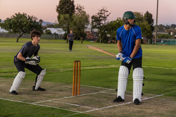 Obraz premium Cricket players male batsman in helmet holding bat, teenage keeper crouching behind stumps on pitch