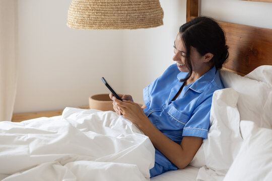 Woman relaxing and smiling on bed with white pillows and wooden headboard, holding smartphone