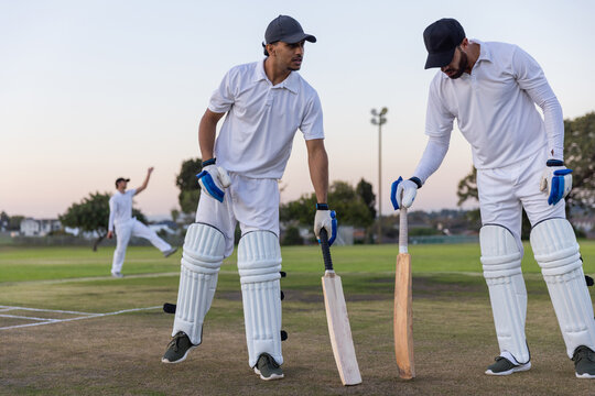 Diverse male teammates standing on cricket pitch in white kit wearing pads and gloves, holding bats