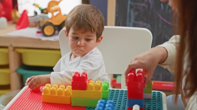 Woman guiding boy's hand placing green plastic block on colorful tower in preschool; focused concentration.