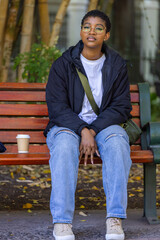Fototapeta premium African American woman sitting on wooden bench in park wearing glasses holding coffee, green bag