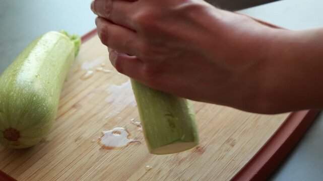 Close-up of a woman's hands using a sharp chef's knife to slice fresh green zucchini on a wooden cutting board in a bright kitchen