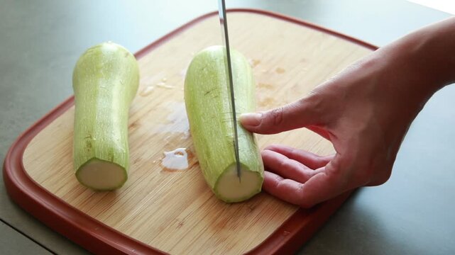 Close-up of a woman's hands using a sharp chef's knife to slice fresh green zucchini on a wooden cutting board in a bright kitchen