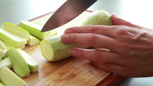 Close-up of a woman's hands using a sharp chef's knife to slice fresh green zucchini on a wooden cutting board in a bright kitchen