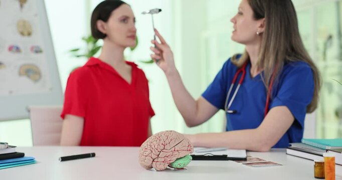 Human brain model and doctor with neurological hammer checks patient reactions in hospital. Physician examines woman reflexes in clinic slow motion