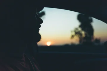 Fototapeten Schwarz Silhouette of bearded man looking through car window at golden sunset during a peaceful evening road trip  © Екатерина Лукашина