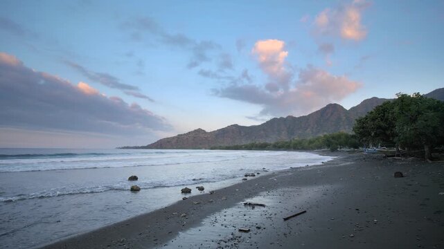 Wide coastal view of Pemuteran Beach, Bali, at dusk with gentle waves washing a dark volcanic shoreline. Mountains and tropical trees frame the bay beneath pastel clouds in soft evening light