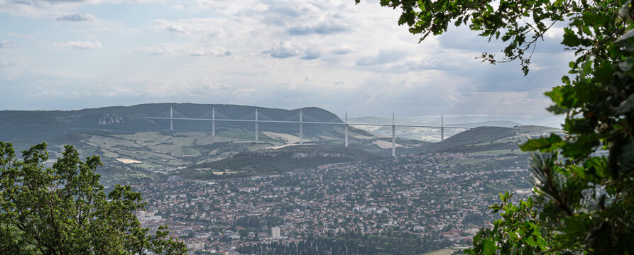 Millau Viaduct rising above the town of Millau in Aveyron, France. Iconic cable stayed bridge spanning the Tarn Valley framed by woodland and dramatic summer clouds.