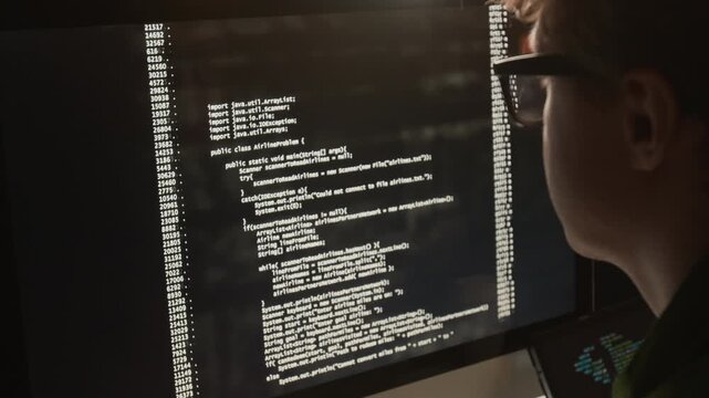 Over the shoulder view of Caucasian young programmer wearing glasses analyzing code on computer screen in dark office. Footage for software development promos and IT education campaigns