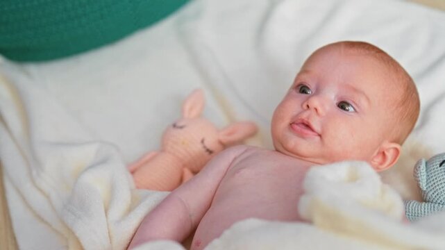 Baby girl clutching cream blanket and pink knitted bunny in a crib, bare face turned upward with hand gripping blanket; serenity.