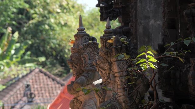Close-up of a traditional Balinese guardian statue at a Hindu temple in Bali, Indonesia. Weathered stone carving adorned with a flower offering stands against lush tropical rainforest