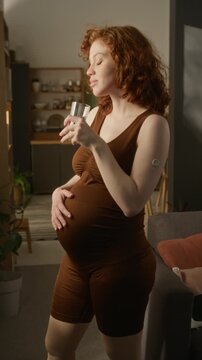 Vertical shot of pregnant Caucasian young adult woman with glucose monitor on arm drinking water and touching belly. Footage suitable for gestational diabetes healthcare marketing, vertical