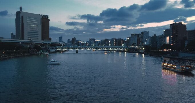 Wide static view, Tokyo cityscape over river in late evening. Tranquil view of Sumida river in Japan downtown. Boats sailing under illuminated bridge. Cityscape lighting up against twilight sky clouds