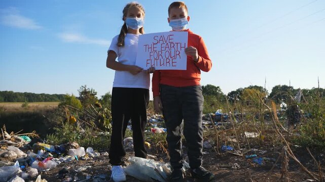 Little boy and girl hold in hands poster of environmental movement for saving earth. Small eco volunteers in protective masks looking into camera and standing against background of garbage. Close up
