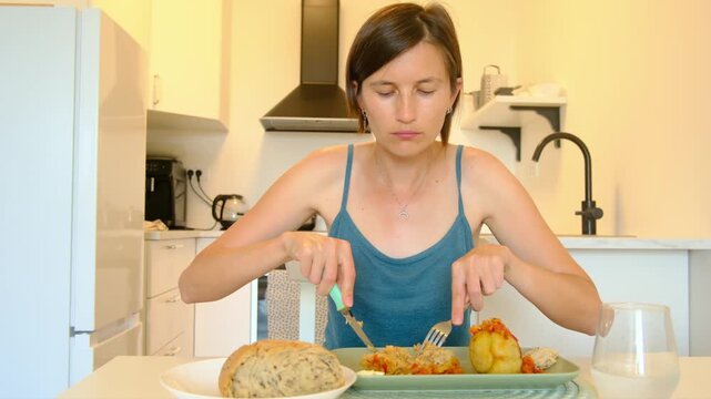 Woman sits at kitchen table eating stuffed paprika, enjoying homemade Balkan cuisine, turning meal into cultural moment shared in quiet domestic space.