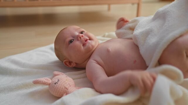 Girl holds soft blanket to mouth while lying on a rug and reaching up in a building nursery room; comfort curiosity.