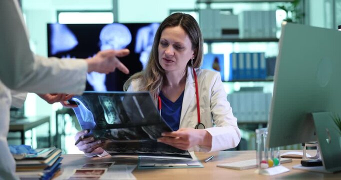 Assistant gives patient spine x-ray image to doctor in clinic. Woman traumatologist examines backbone scans with female colleague in hospital