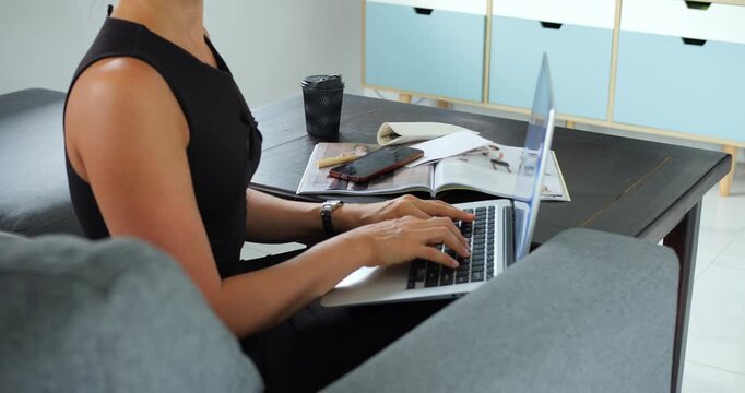 Hands typing on laptop from seated position, businesswoman in black dress working on planning and spreadsheets phone, documents and takeaway cup on table create organized home office atmosphere.