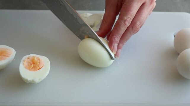 Close-up of a woman's hands cleanly slicing a hard-boiled egg in half with a sharp knife on a white cutting board