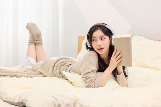 Young woman relaxing on bed with headphones and book at home