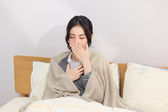 Sick young woman sitting in bed wrapped in blanket and feeling nauseous