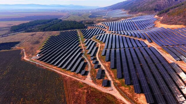 Large solar park with valley and town in background, Bulgaria. Wide drone perspective of multi-section panel fields, red soil, vineyard rows and distant mountain range.