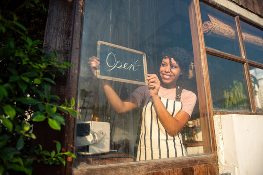 Person in apron holding 'Open' sign inside a rustic cafe window.