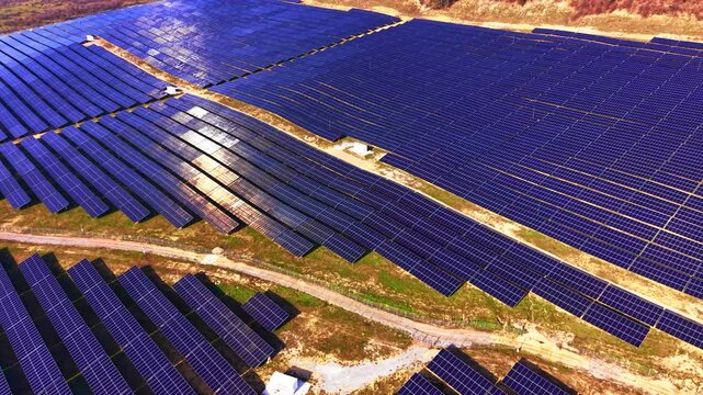 Solar farm sections divided by access roads, Bulgaria. Elevated drone view of blue panel blocks separated by wide dirt paths, small utility buildings and green grass verges.