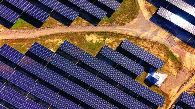 Solar panels beside curved gravel road on hillside. Top-down drone view showing winding grey gravel track separating panel array from green and brown scrubland.