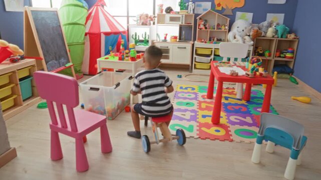 Little boy rides wooden tricycle, grips handlebars and pushes pedals in a bright playroom building full of toys and blocks; playful childhood joy.