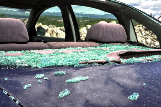 Detailed view of a dark blue sedan with a completely shattered, imploded tempered rear window. Shards are scattered across the trunk and parcel shelf. Mountains in the distance.