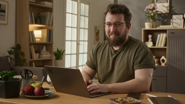 Portrait of middle aged Caucasian man with diabetes CGM sensor on arm typing on laptop at home and smiling. Footage ideal for diabetes awareness campaigns and digital health marketing