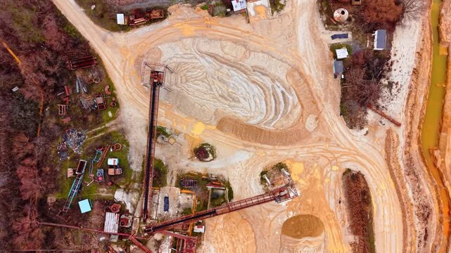 Overhead view of conveyor belt and sand stockpile at quarry site, Bulgaria. Rusty industrial equipment, white sand mound, orange-stained ground and scattered machinery.