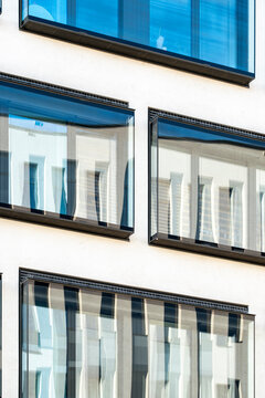 The windows repeat across a white facade as glass reflection catches the urban building in blue daylight, while curtains and shadow soften the exterior rhythm