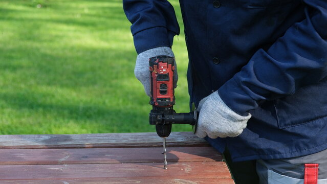 A close-up of a worker in a blue jacket and protective gloves drilling a hole in a wooden table outdoors with a cordless impact drill. Construction, carpentry, and home renovation concept. Banner