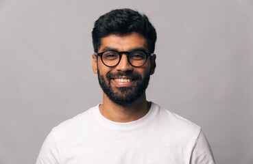 Obraz premium Portrait of a Smiling Young Indian Man with Glasses and Beard Wearing a White T-shirt Against a Neutral Background