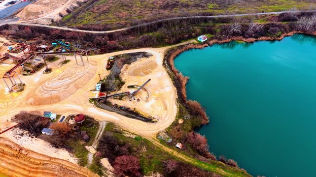 Elevated view of sand processing plant beside turquoise quarry lake. Oblique drone angle showing conveyor towers, sand piles, reed-lined shore and vivid teal water under winter light.