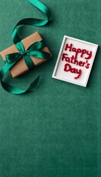 Minimalist flat lay composition for Father's Day with a small brown gift box tied with emerald green ribbon and "Happy Father's Day" framed text on a deep green textured surface.
