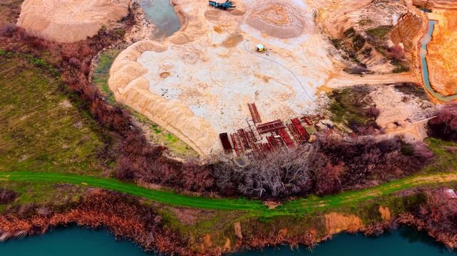Drone view of quarry lake shore with stacked steel frames. Oblique perspective of red steel structure piles on sandy peninsula, teal lake water, green strip and bare deciduous trees.