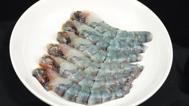 Freshly peeled and headless black tiger shrimp neatly arranged on a white plate, slowly rotating against a black background.