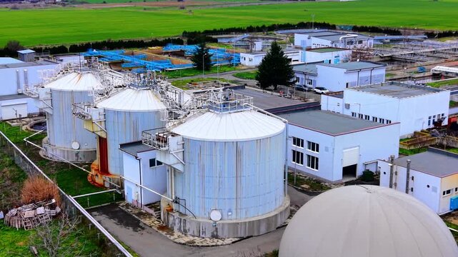 Elevated view of wastewater treatment plant digester tanks. Oblique drone view large corrugated metal silos, pipework, dome structure, white buildings and green winter fields.