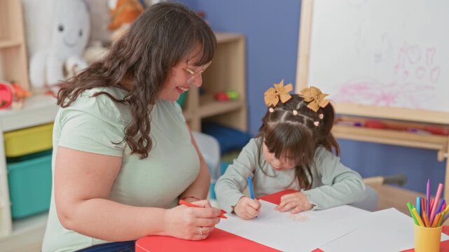 Woman guides girl's hand as she draws with a blue marker on paper at a red table, pencils and crayons nearby and a whiteboard with scribbles in a classroom building; nurturing learning focus.