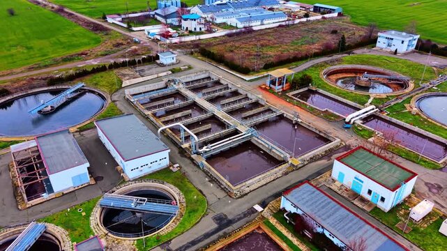 Aerial view of full wastewater treatment complex near agricultural fields. Wide drone shot showing clarifiers, aeration basins, buildings, green winter fields and overcast sky above.
