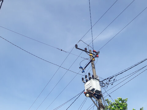 A low angle view of an electrical distribution transformer mounted on a utility pole with power lines and cables above it, against a clear blue sky.